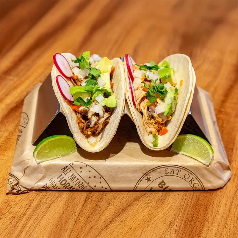 Two vibrant tacos with meat and fresh toppings in a metal holder.