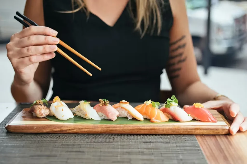 Various nigiri sushi on wooden board with hands holding chopsticks.