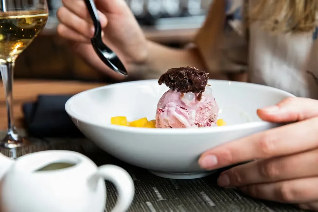 Pink ice cream with chocolate, fruit, and a drink, held by hands.