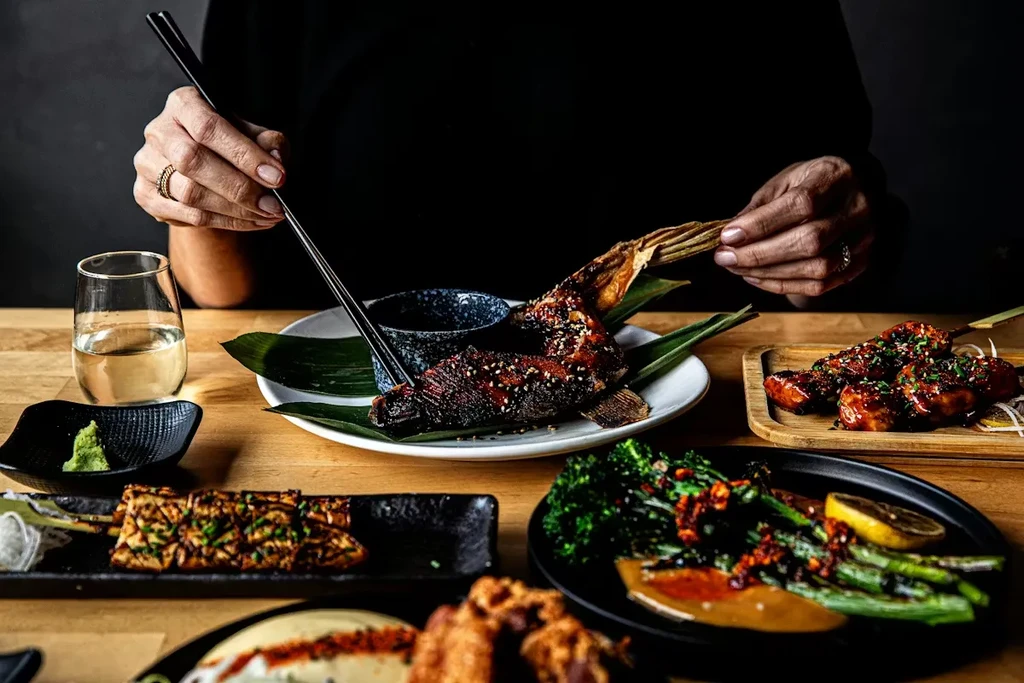 Close-up of hands picking grilled fish with chopsticks from a laden table.