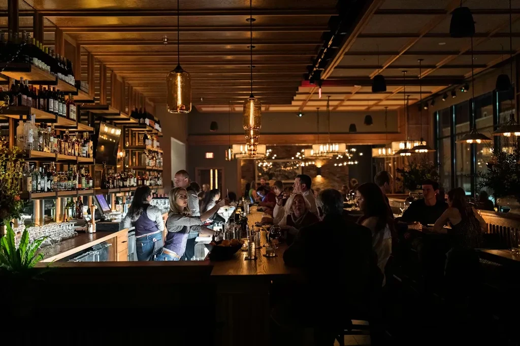 Lively bar interior with bartenders, patrons, warm lighting, and wood.