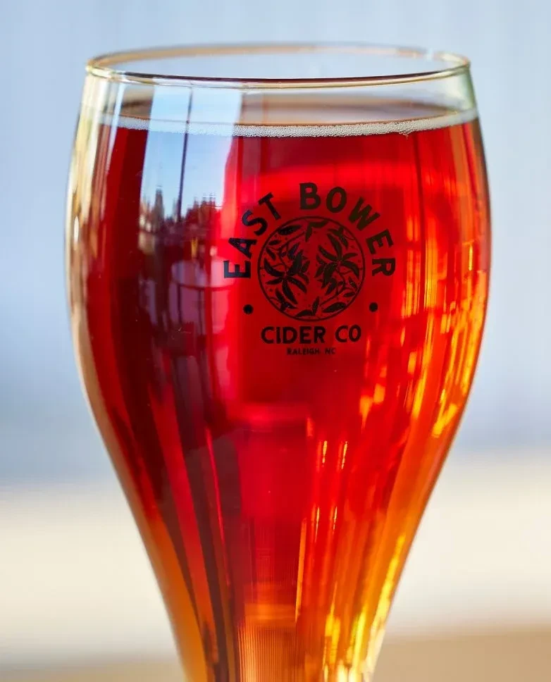 Reddish-orange cider in a stemmed glass with foam and East Bower logo.