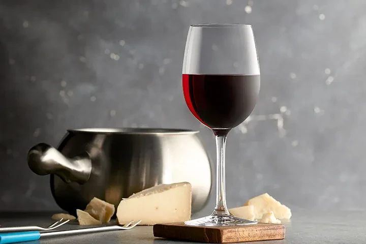Cheese fondue pot, wine glass, and cheese with forks on dark background.