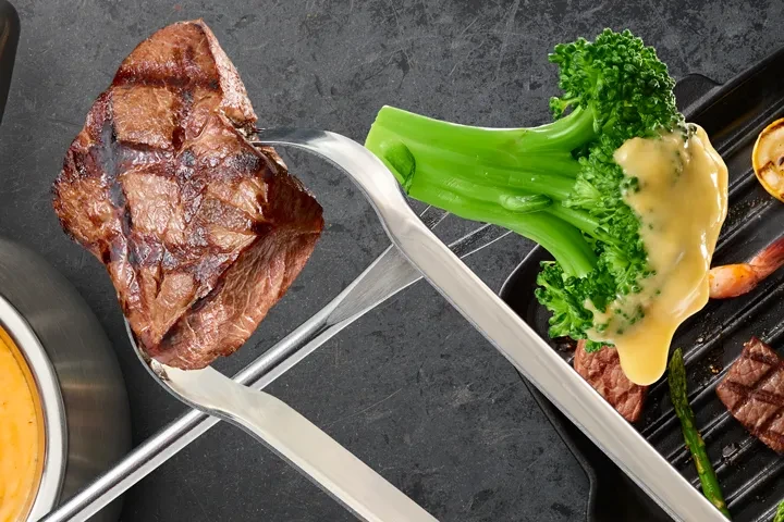 Tongs hold grilled steak and sauced broccoli above a grill pan.