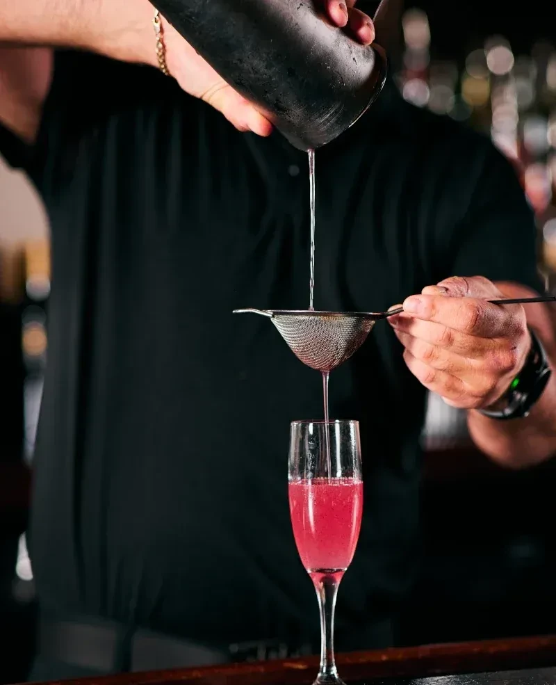 Bartender's hands straining a drink into a flute with pink liquid.