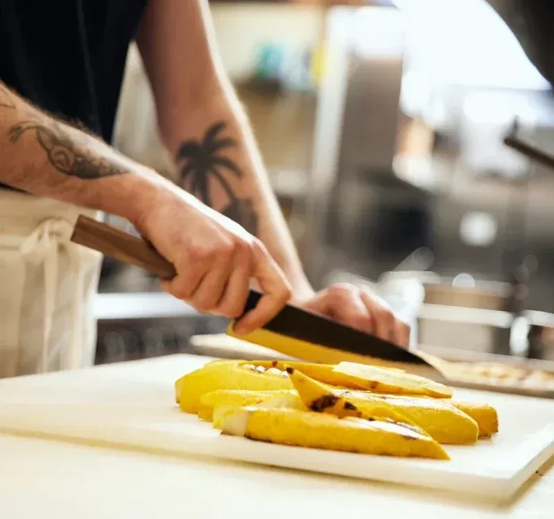 Tattooed hands slicing yellow fruit with a knife in a commercial kitchen.