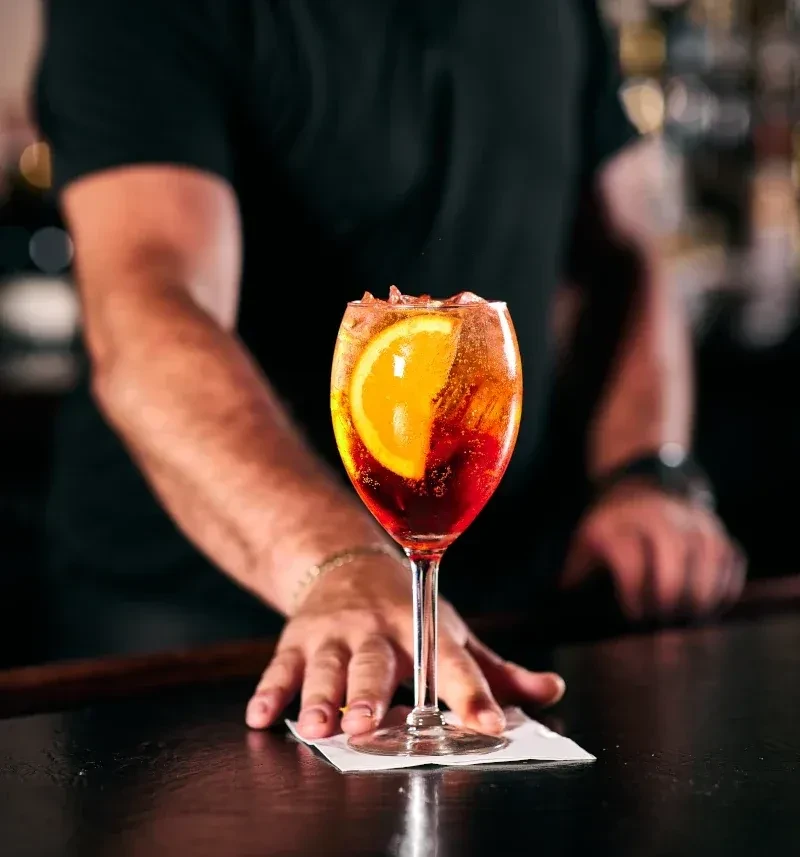 Orange-red cocktail, ice, orange slice on bar; person's hands visible.