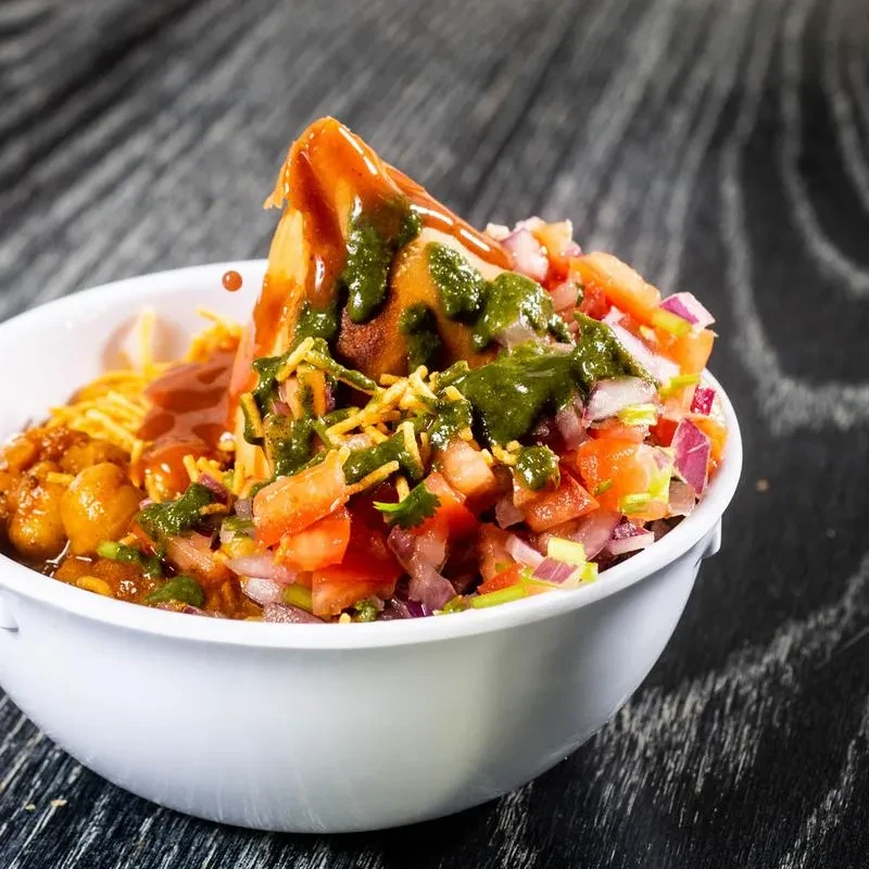 Samosa Chaat in white bowl, topped with chutneys, sev, and vegetables.