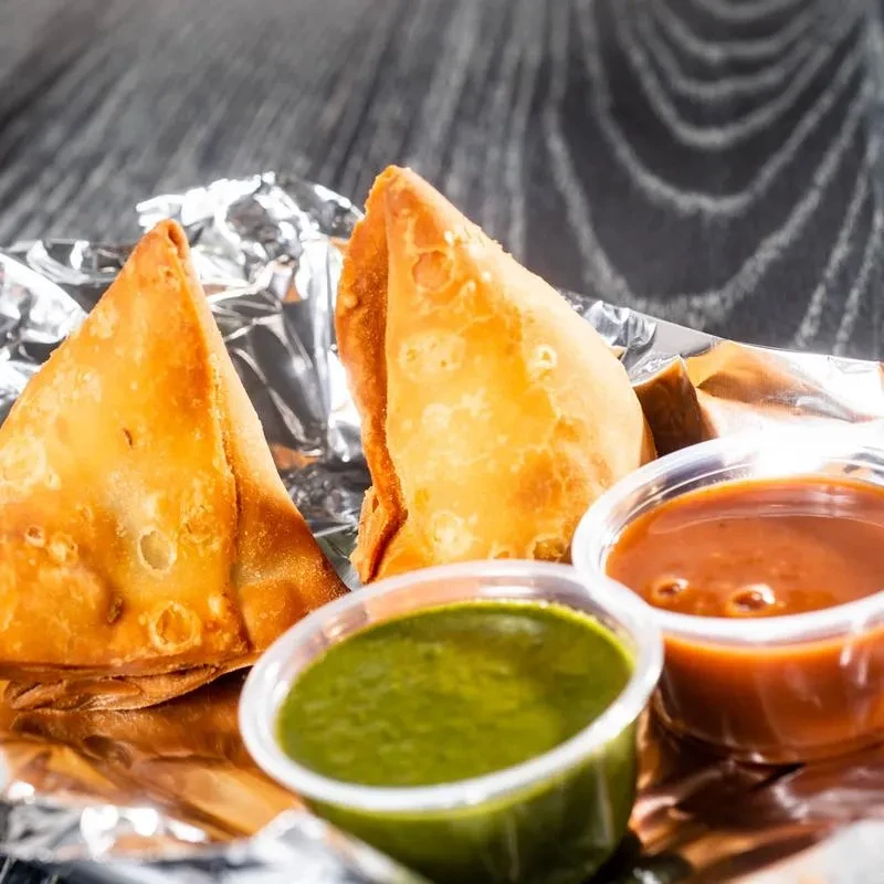 Two crispy fried samosas with two chutneys on aluminum foil.