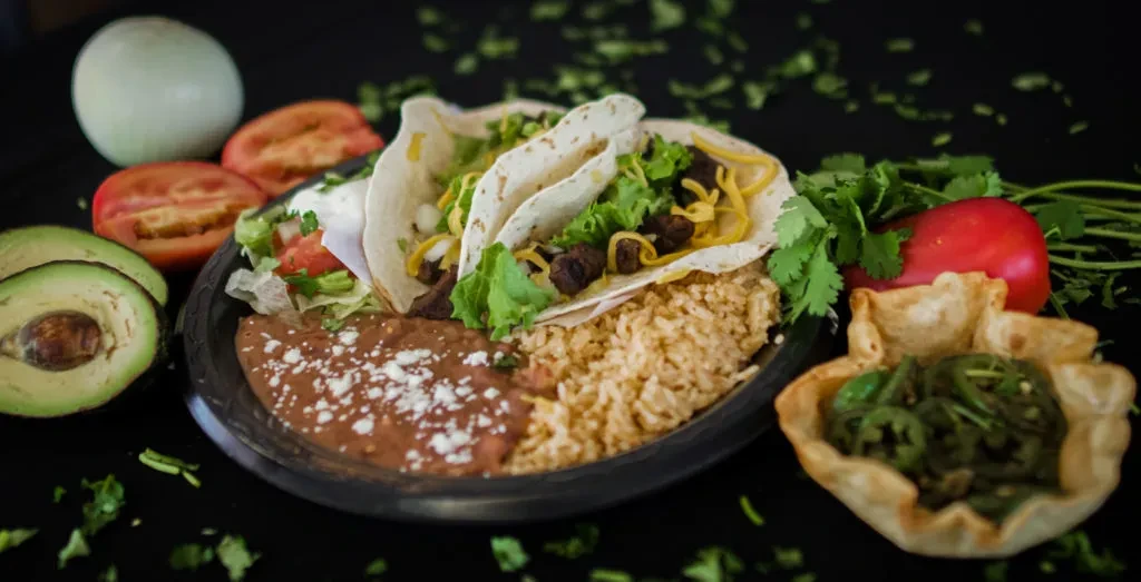 Mexican tacos, rice, refried beans, and fresh produce on dark plate.