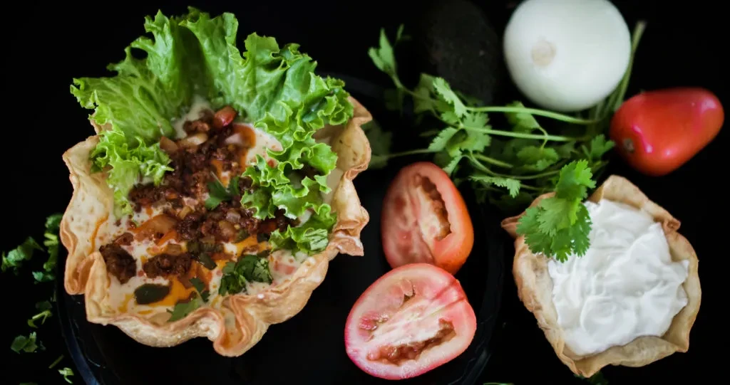 Fried tortilla bowl taco salad with surrounding fresh ingredients.