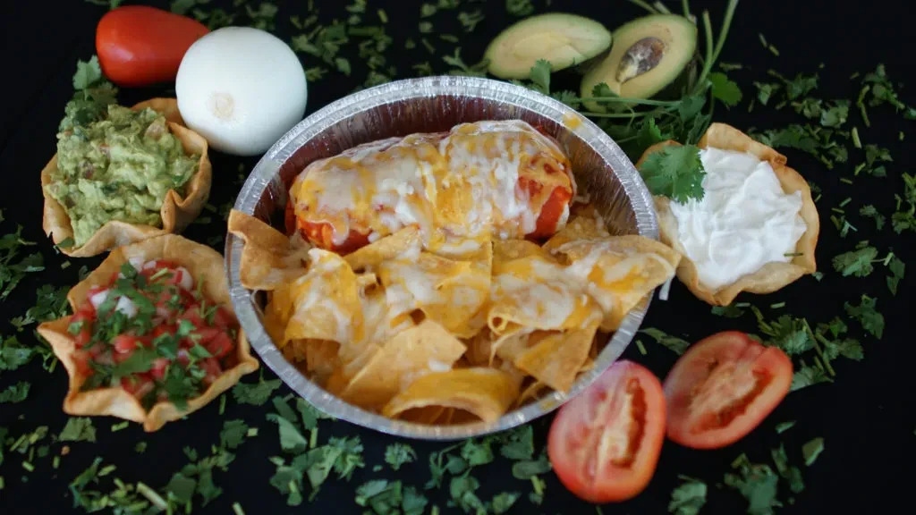 Overhead of cheesy Mexican burrito, chips, guacamole, pico, and fresh ingredients.