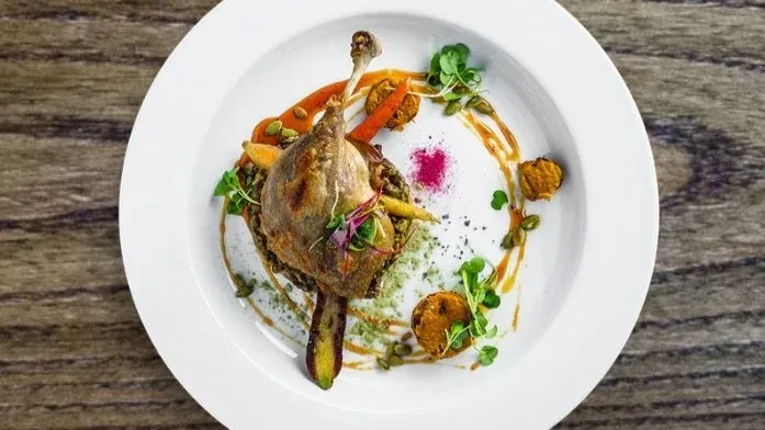 Gourmet plated duck leg confit with grains, carrots, sauces, and microgreens.