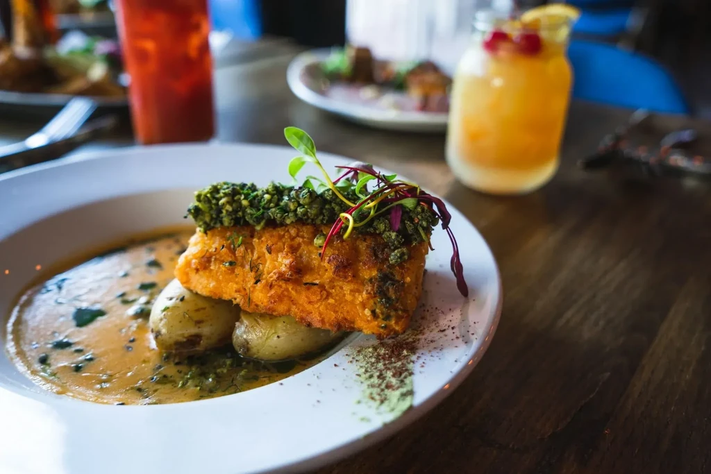 Plated fish with green crust, potatoes, sauce, and microgreens.