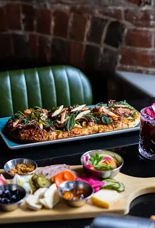 A rustic tabletop features flatbread with meat, charcuterie, and a cocktail.