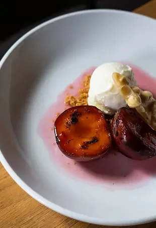 Plated dessert: roasted plums, white ice cream, red sauce, and crumbs.