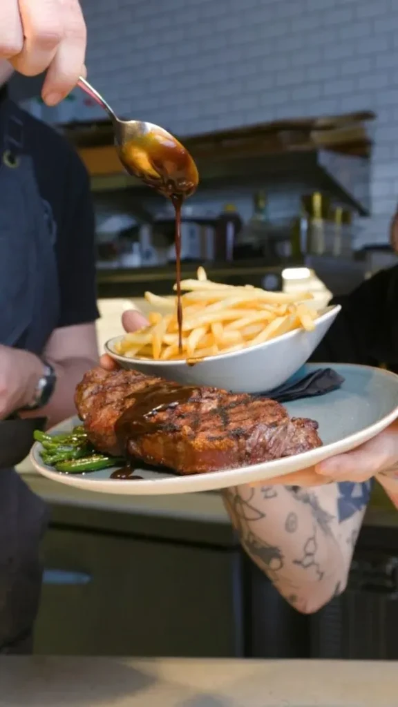 Hand drizzles dark sauce over grilled steak, green beans, and fries.