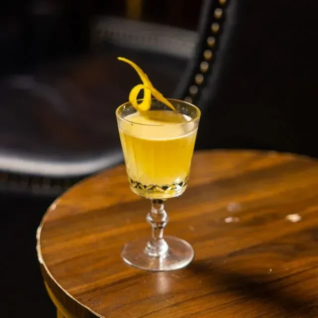 A stemmed glass cocktail with a curled citrus peel on a wood table.