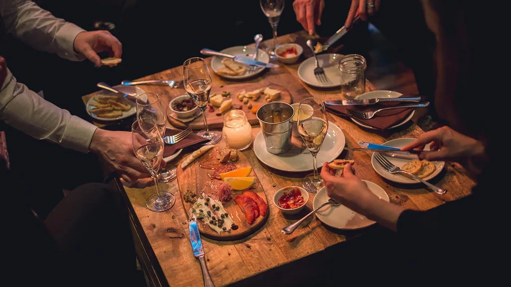 Hands dine on charcuterie and drinks at a cozy, rustic table.