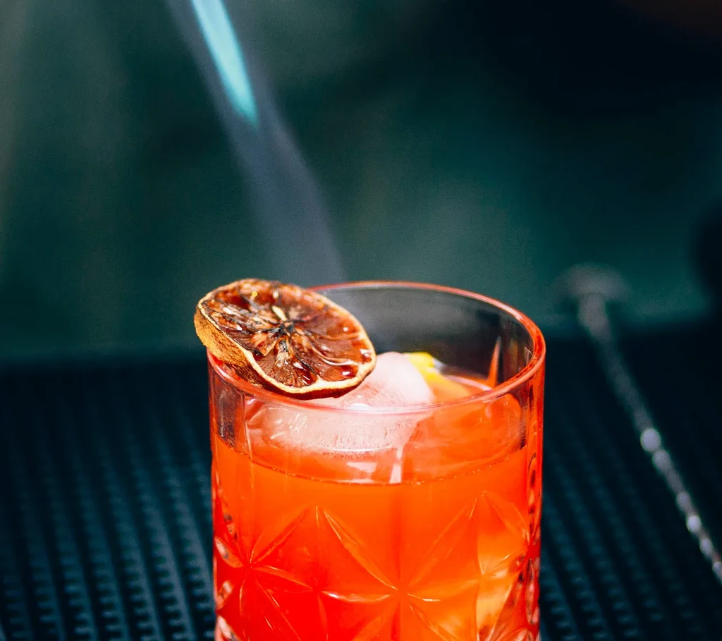 Red cocktail with ice, citrus garnish, and a blue flame.