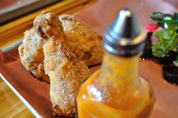 Two fried chicken pieces, hot sauce bottle, and blurred vegetables.