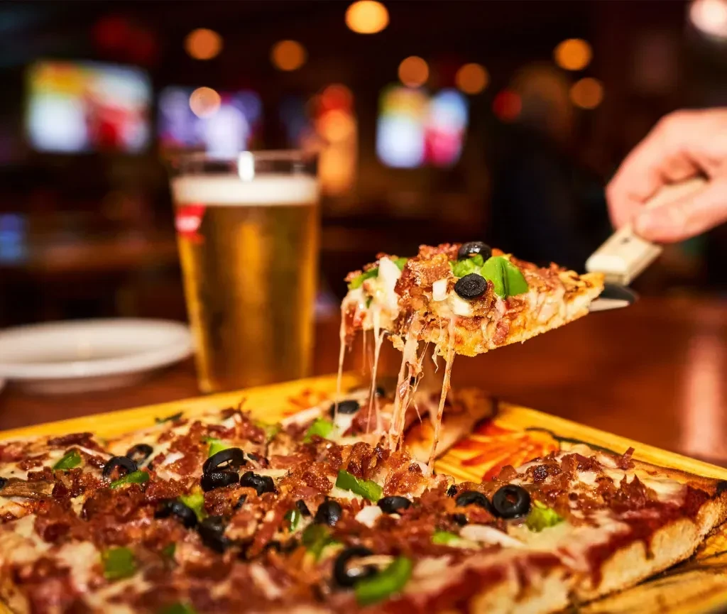 Hand lifts cheesy rectangular pizza with bacon, olives, peppers and beer.
