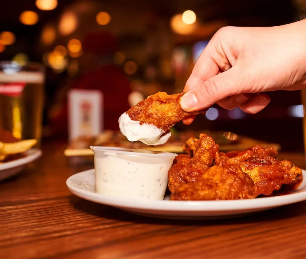 Hand dipping a crispy fried chicken wing into creamy white sauce.