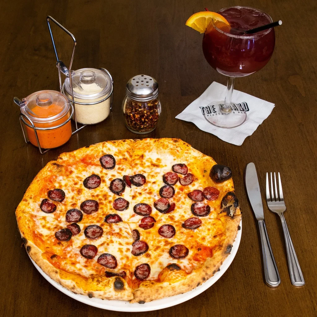 Pepperoni pizza with drink, condiments, and cutlery on a wooden table.