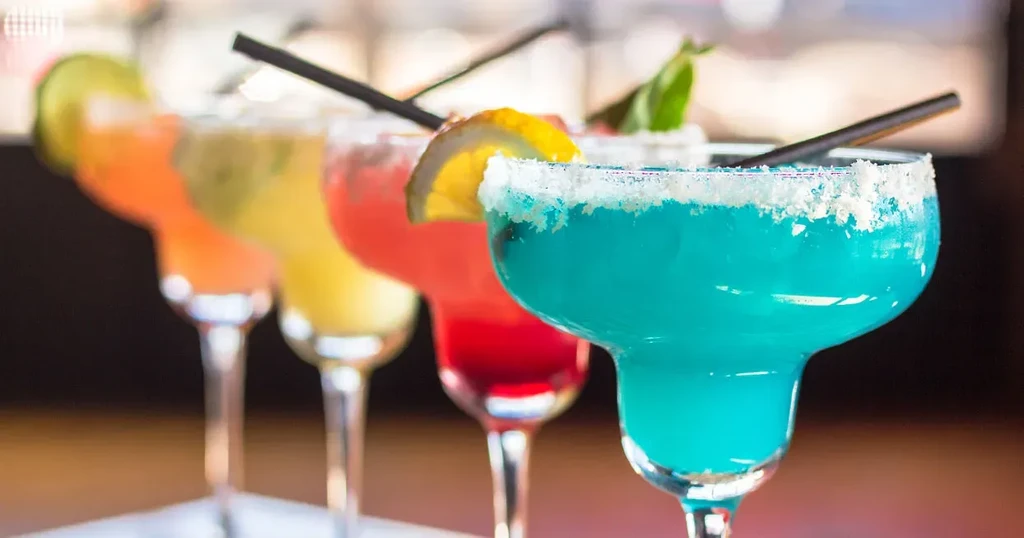 Five colorful margaritas in a row with salted rims and garnishes.