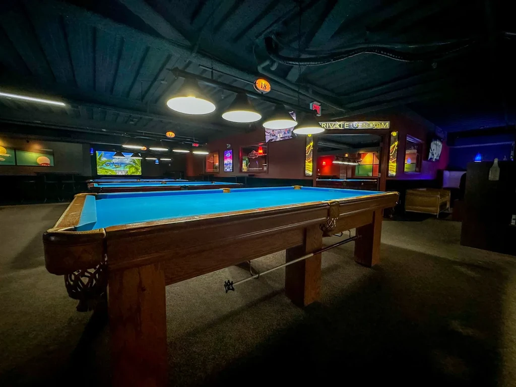 Empty, dimly lit pool hall with multiple blue-felt tables, brightly lit.