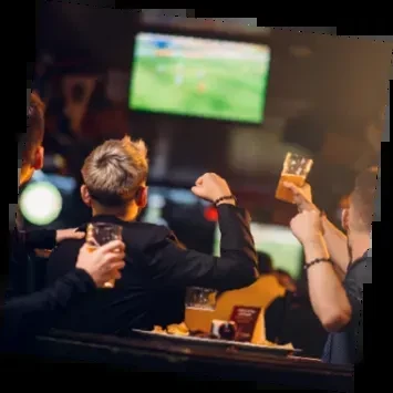 Two men cheer with raised drinks, watching sports in a dark bar.