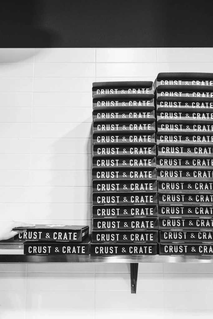 Stacks of "CRUST & CRATE" pizza boxes on a shelf, a hand visible.