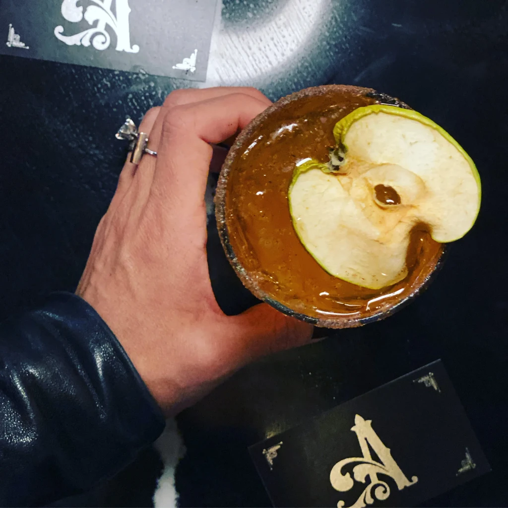 Dimly lit hand with ring holds amber apple cocktail with spiced rim.
