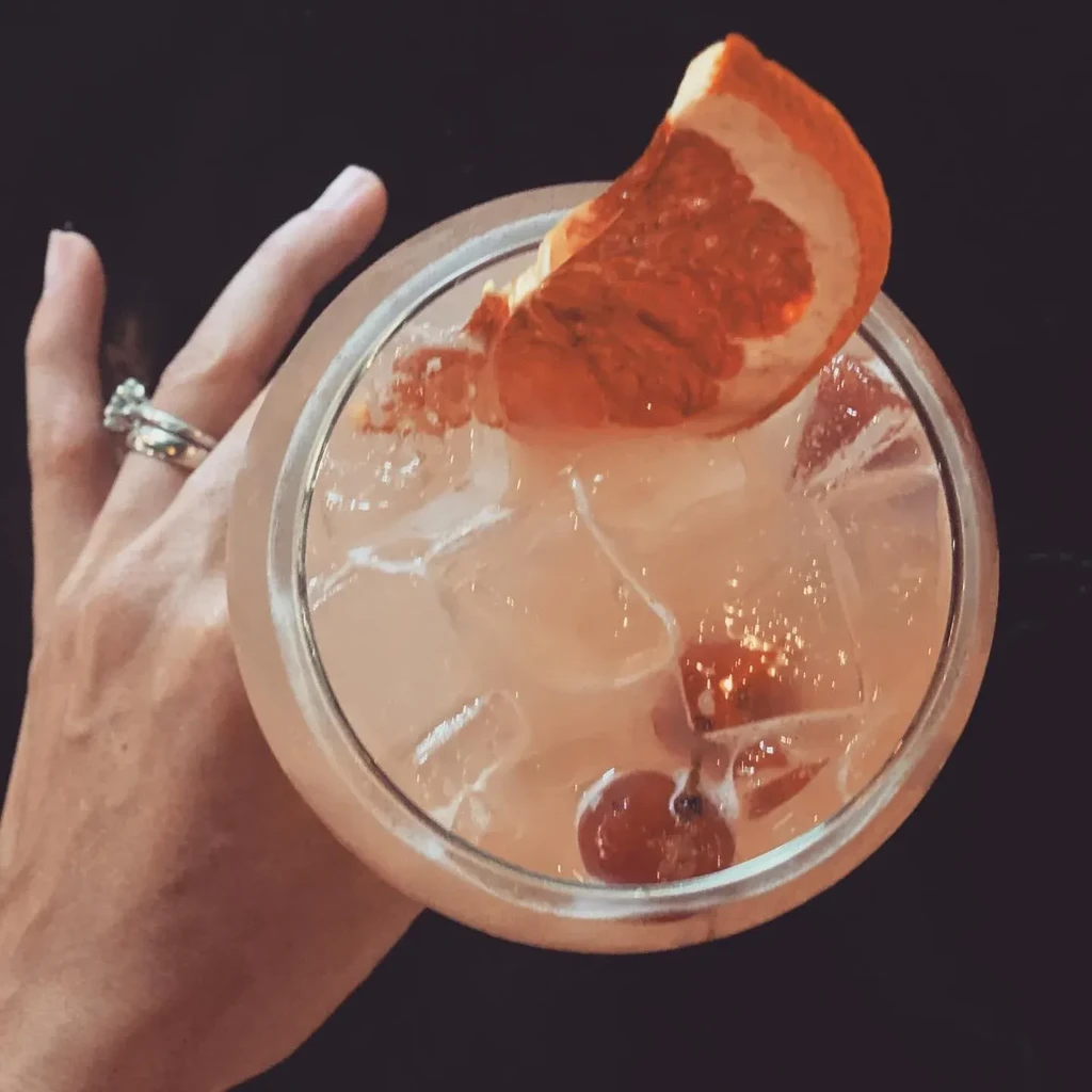 Close-up pink cocktail with ice, dried citrus, berries, and ringed hand.