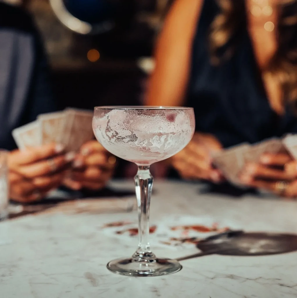 Empty cocktail glass on table, blurred background shows people playing cards.