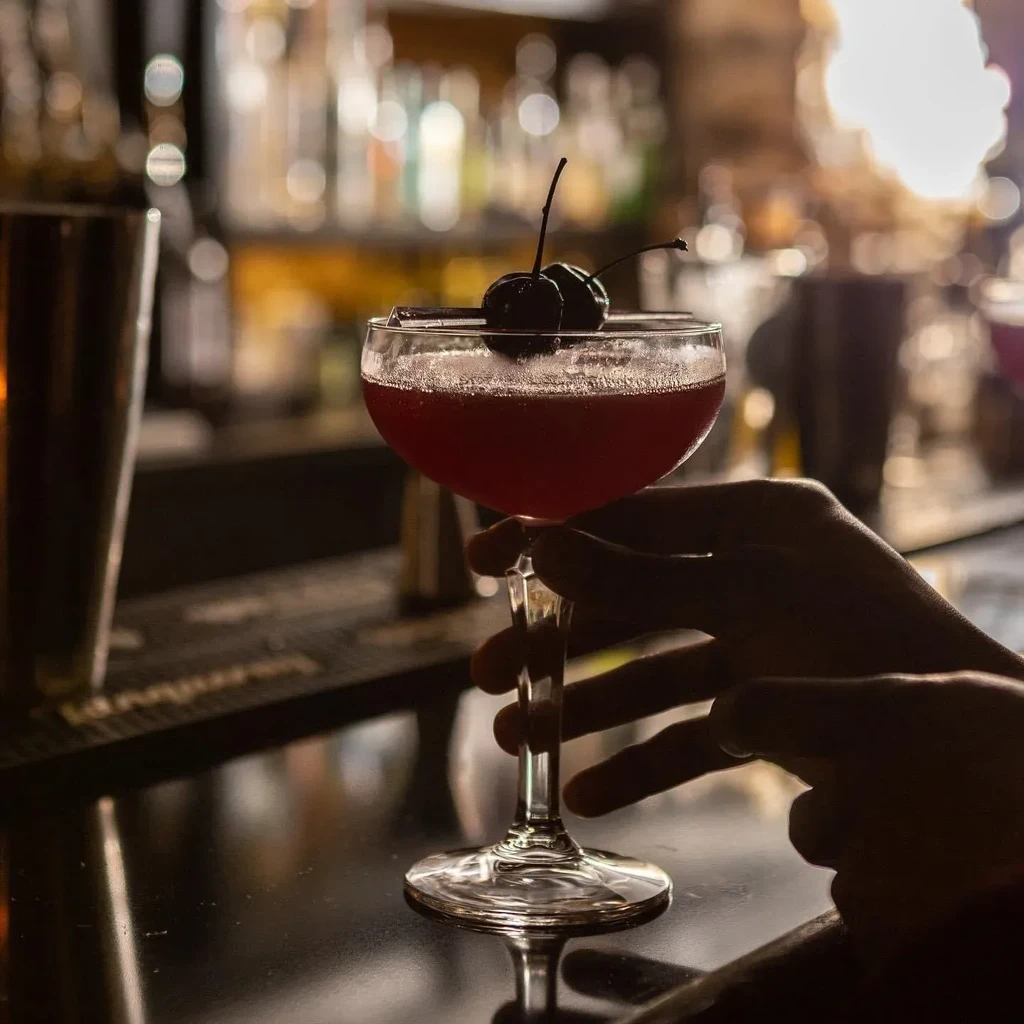 Red cocktail in coupe glass with cherries, held by a silhouetted hand.