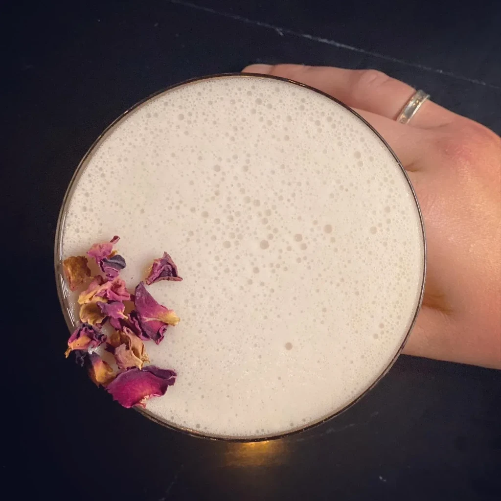 Top-down foamy cocktail with dried rose petals, held by a hand.