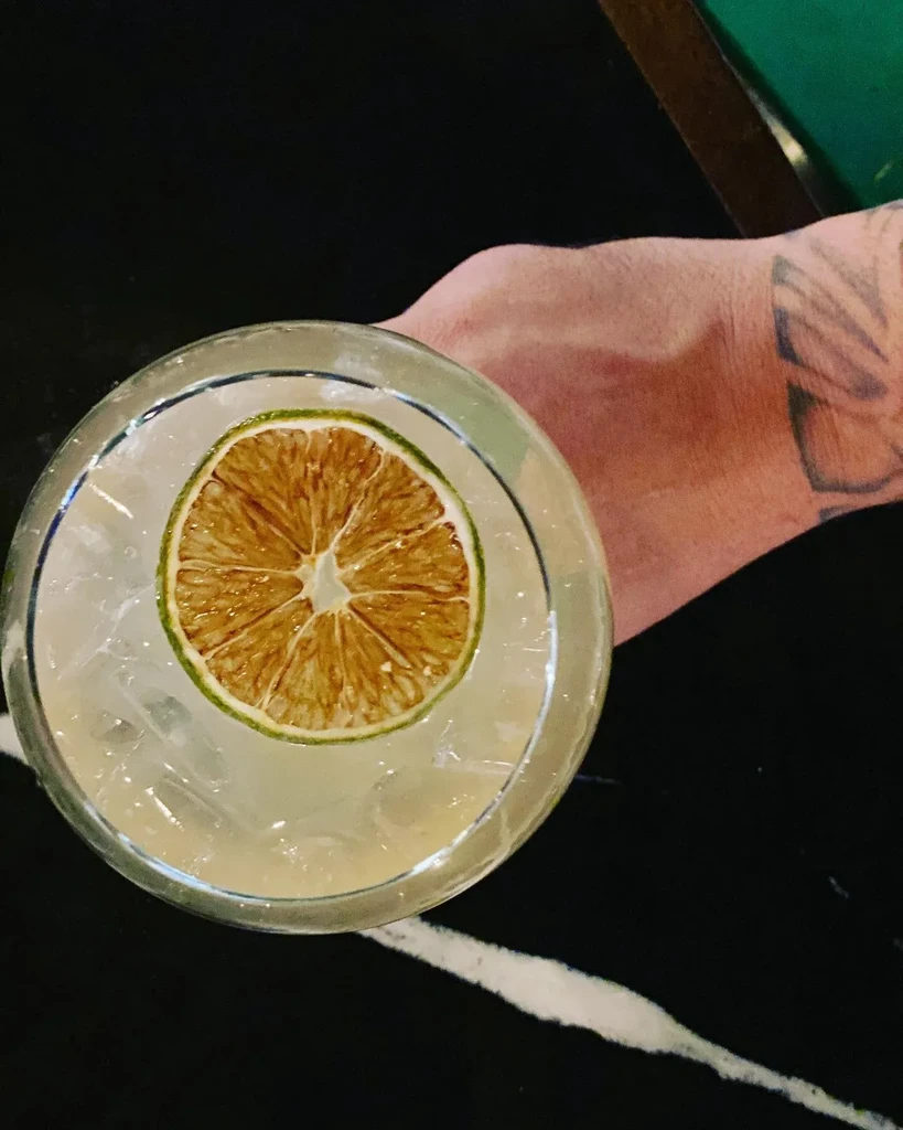 Overhead: Cloudy yellow drink with dried lime, ice, and tattooed hand.