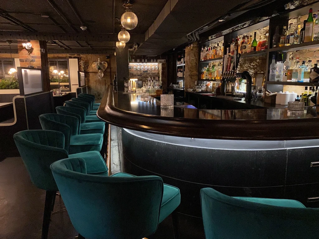Dimly lit bar with curved counter, liquor bottles, and teal stools.