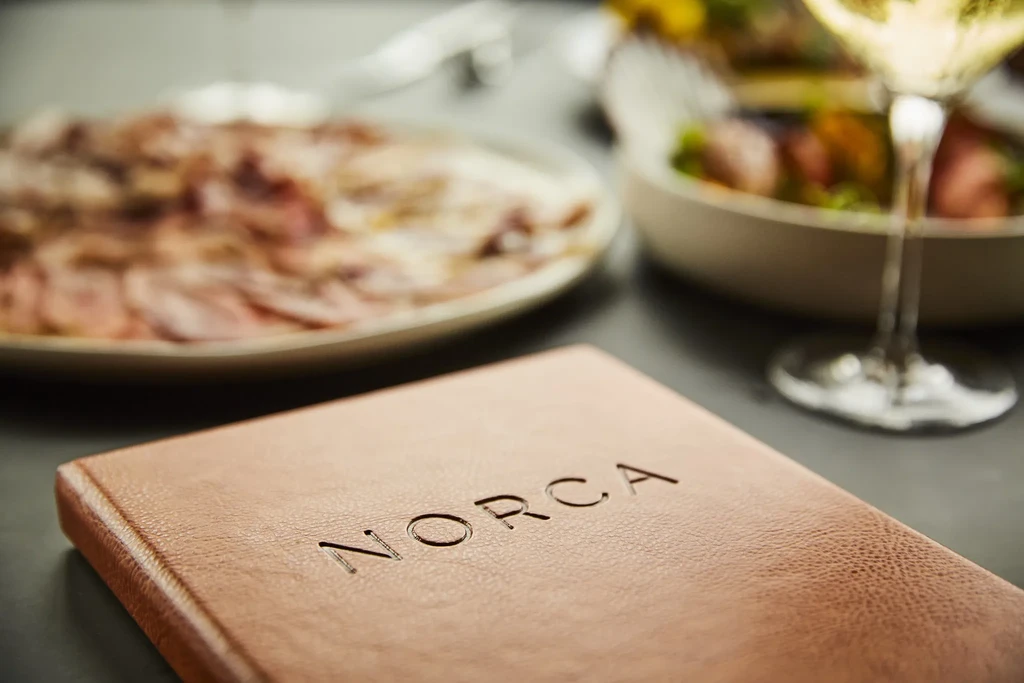 Brown leather "NORCA" menu with blurry fine dining food and wine.