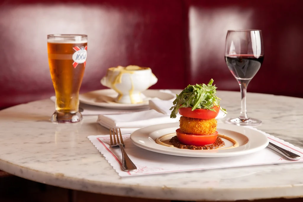 Fried item between tomatoes with arugula, beer, and wine on marble table.