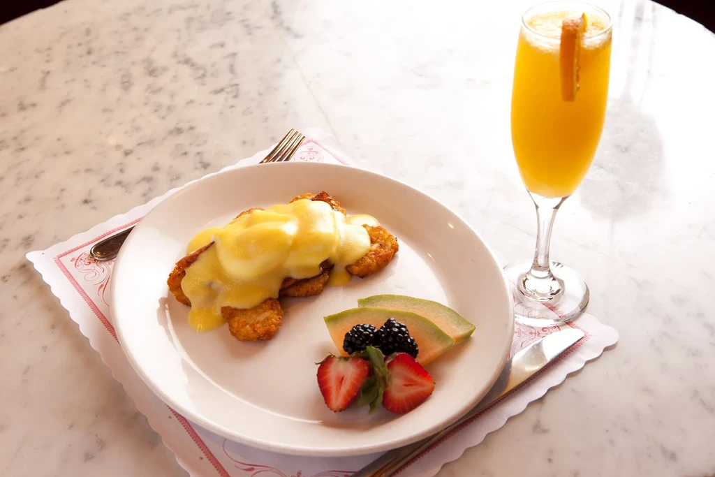 Brunch meal: Eggs Benedict, fresh fruit, and mimosa on a marble table.