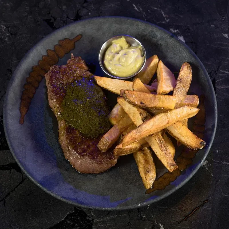 Grilled steak with herb sauce, fries, and dipping sauce on dark plate.