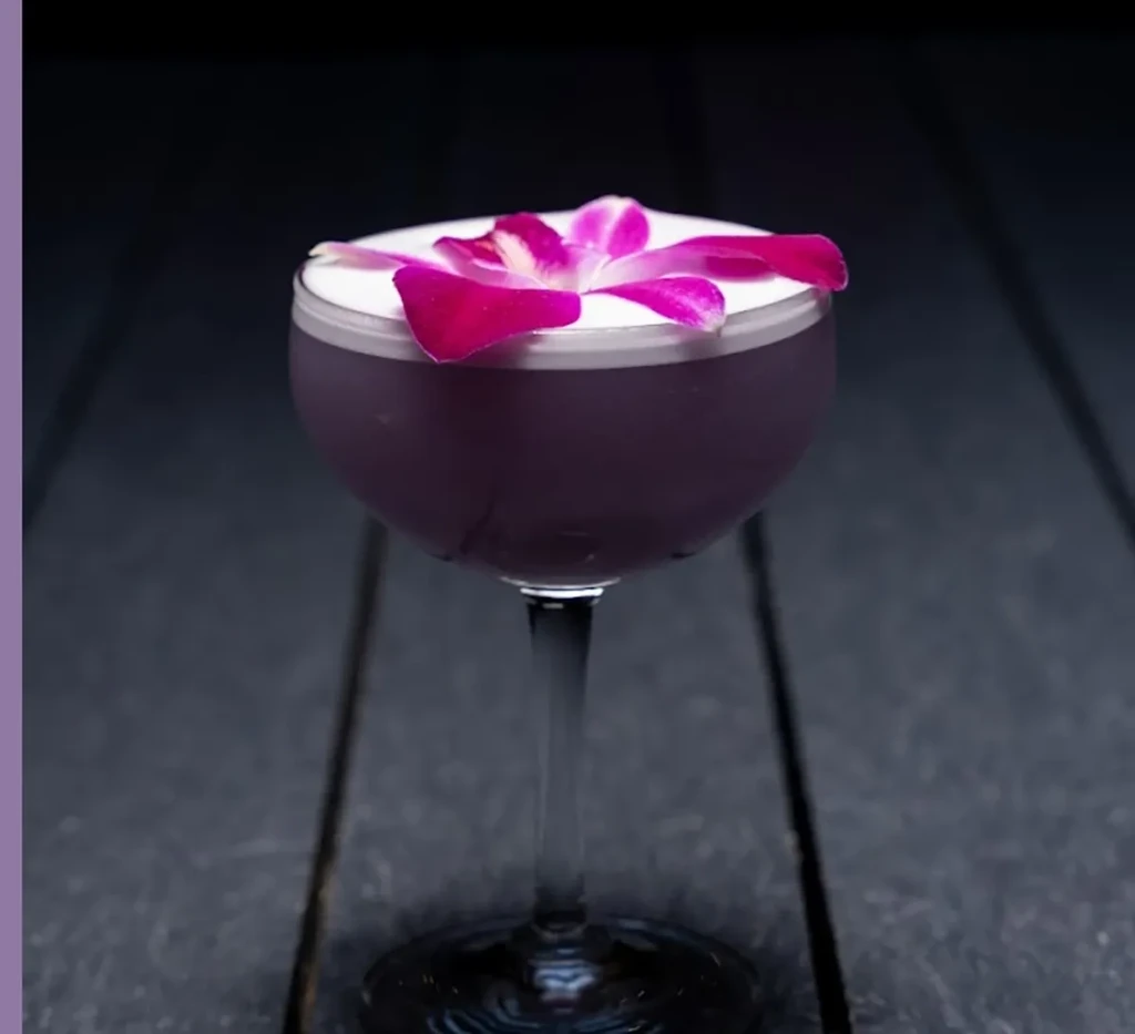 Purple cocktail in a coupe glass with white foam and orchid garnish.