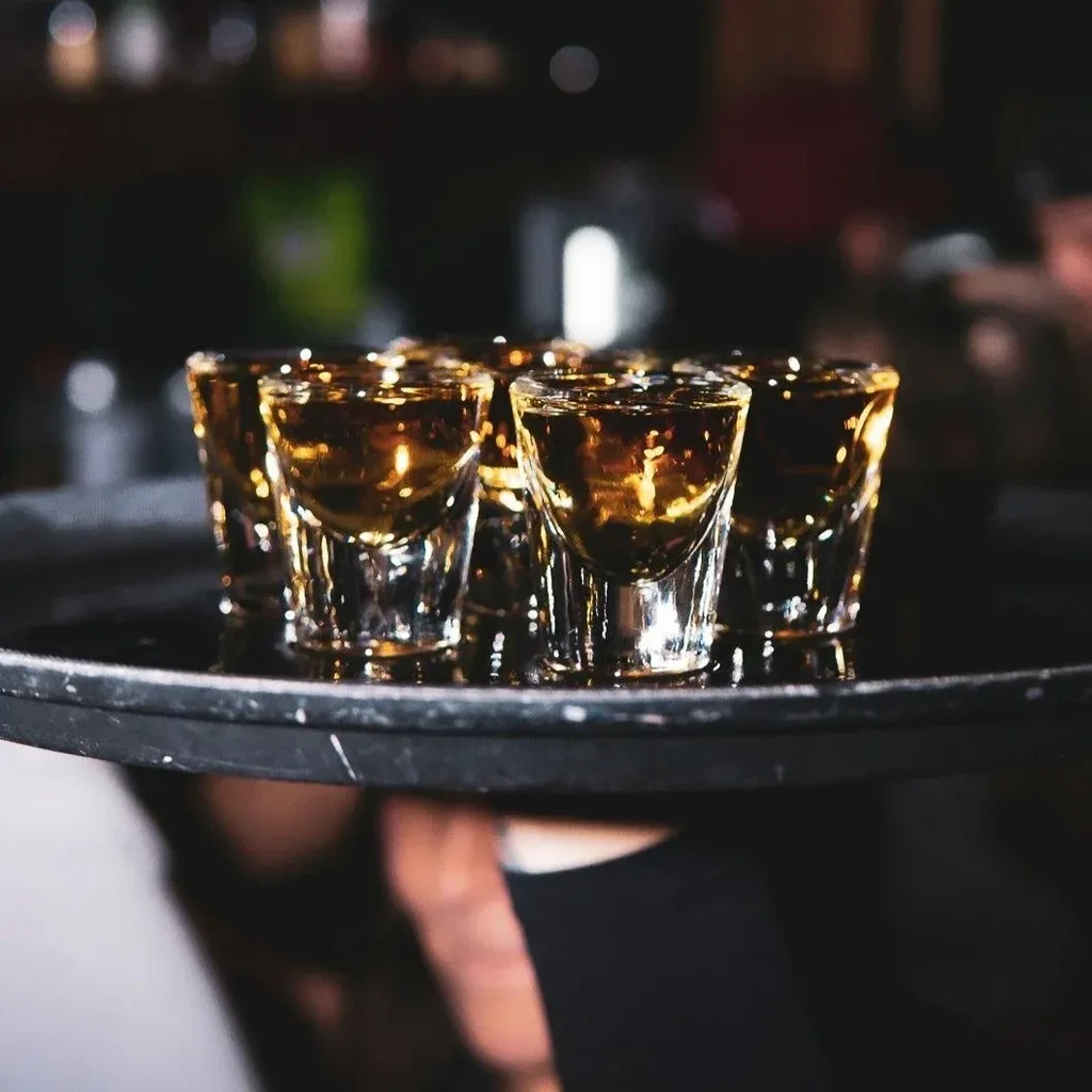 Six shot glasses of amber spirits on a dark tray.
