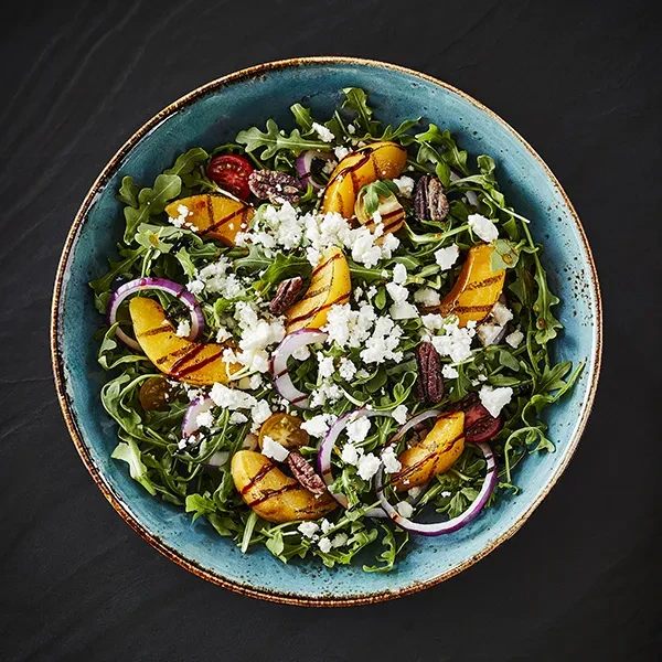 Grilled peach arugula salad with cheese, pecans, tomatoes, teal bowl.