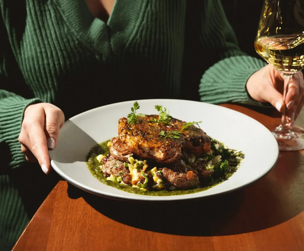 Plated pan-seared meat, smashed vegetables, green sauce, and white wine.