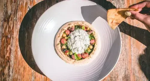 Hummus dish with tomatoes, chickpeas, herbs, and a hand holding pita.
