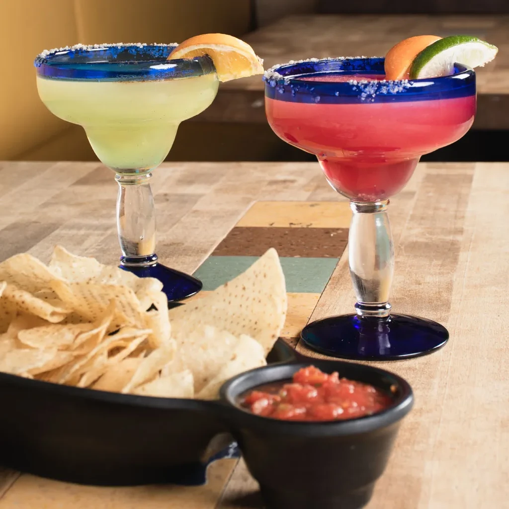Two blue-rimmed margaritas, tortilla chips, and salsa on a striped table.