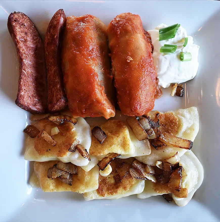 Platter of Eastern European food: kielbasa, golabki, pierogi, sour cream, onions.